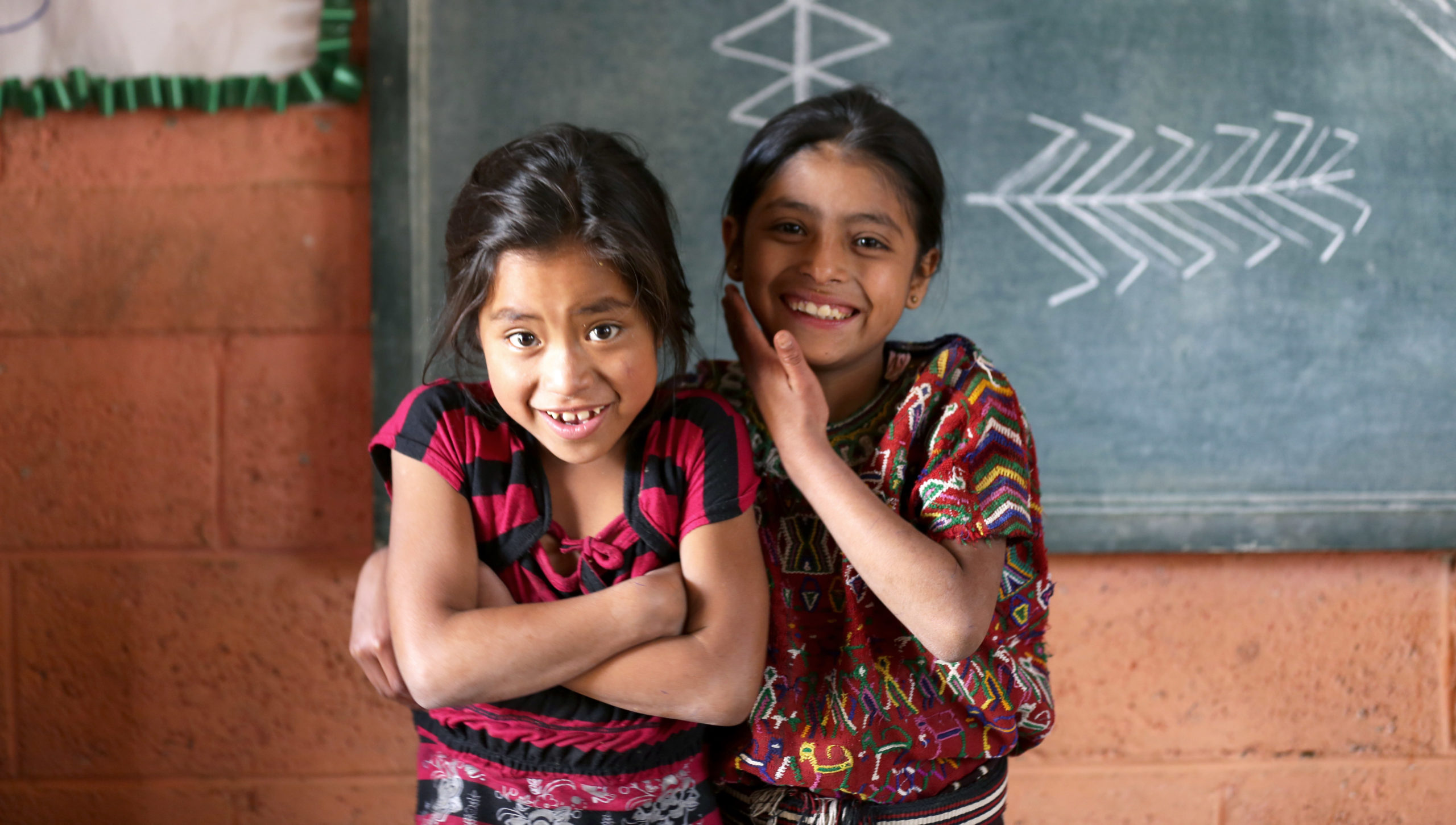 Two students in Guatemala stand in front of a blackboard inside a classroom.